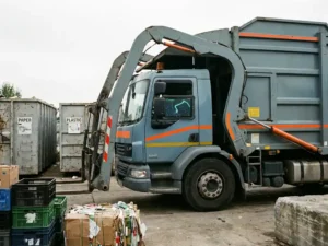 Müllentsorgungsfahrzeug mit Hydraulikarmen an Recyclinghof, digitales Routenoptimierungstablet im Führerhaus sichtbar